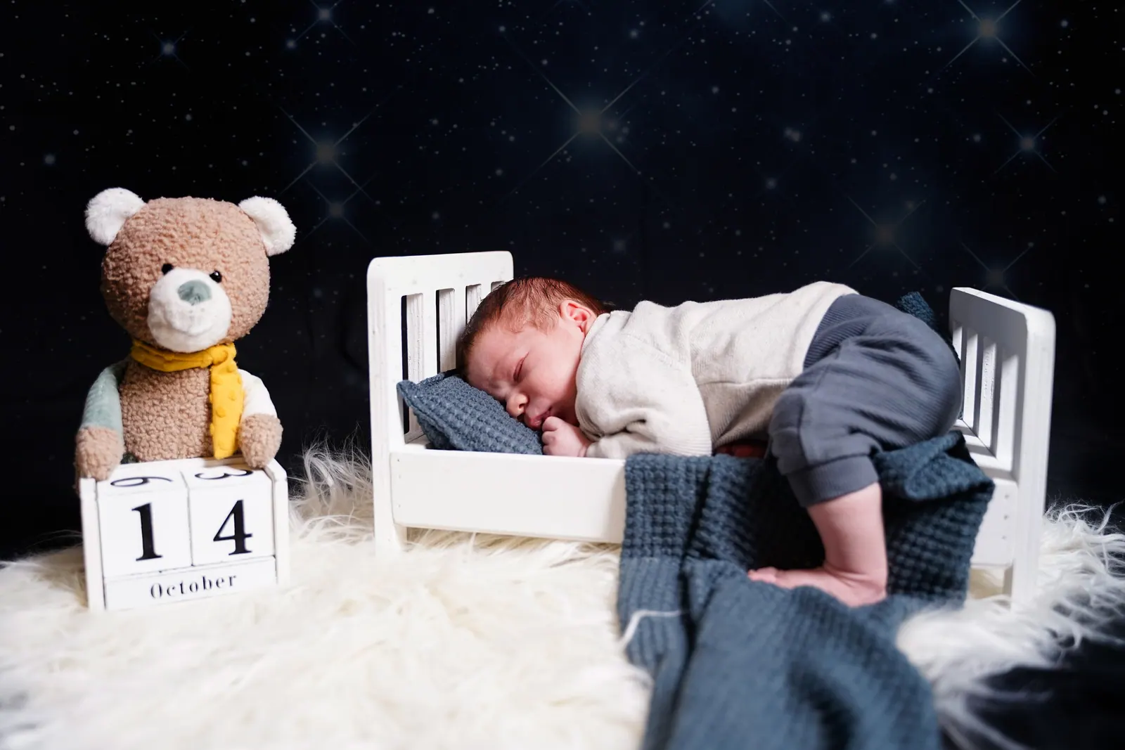 Ein Teddybär mit einem Holzkalender sitzt neben dem kleinen weißen Bettchen, in dem ein Neugeborenes vor sternenklarem Hintergrund schläft