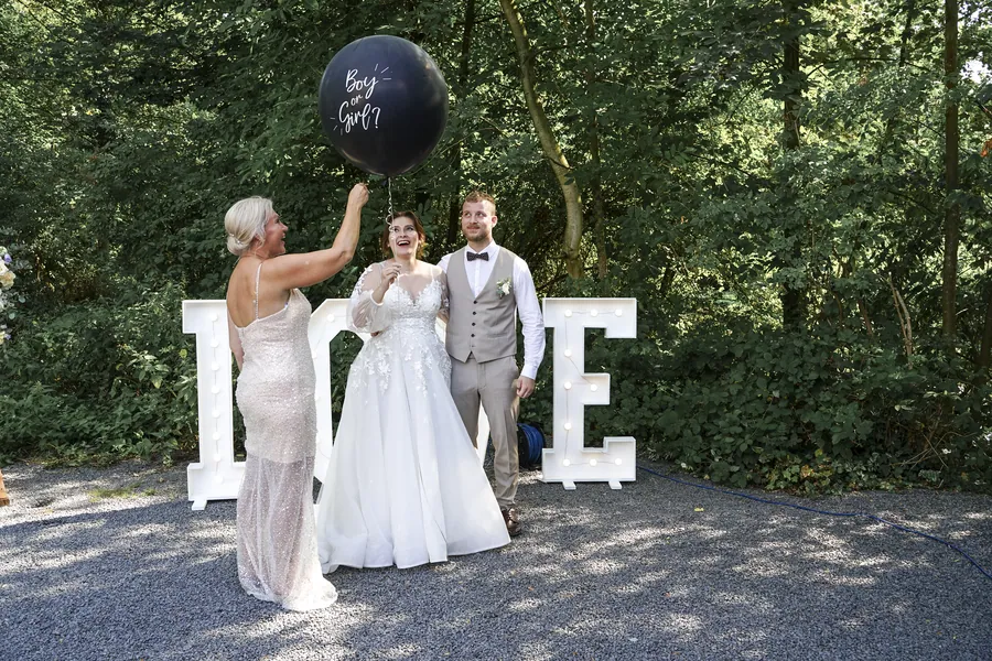 Drei festlich gekleidete Personen stehen mit einem schwarzen Ballon vor großen LOVE-Buchstaben im Grünen