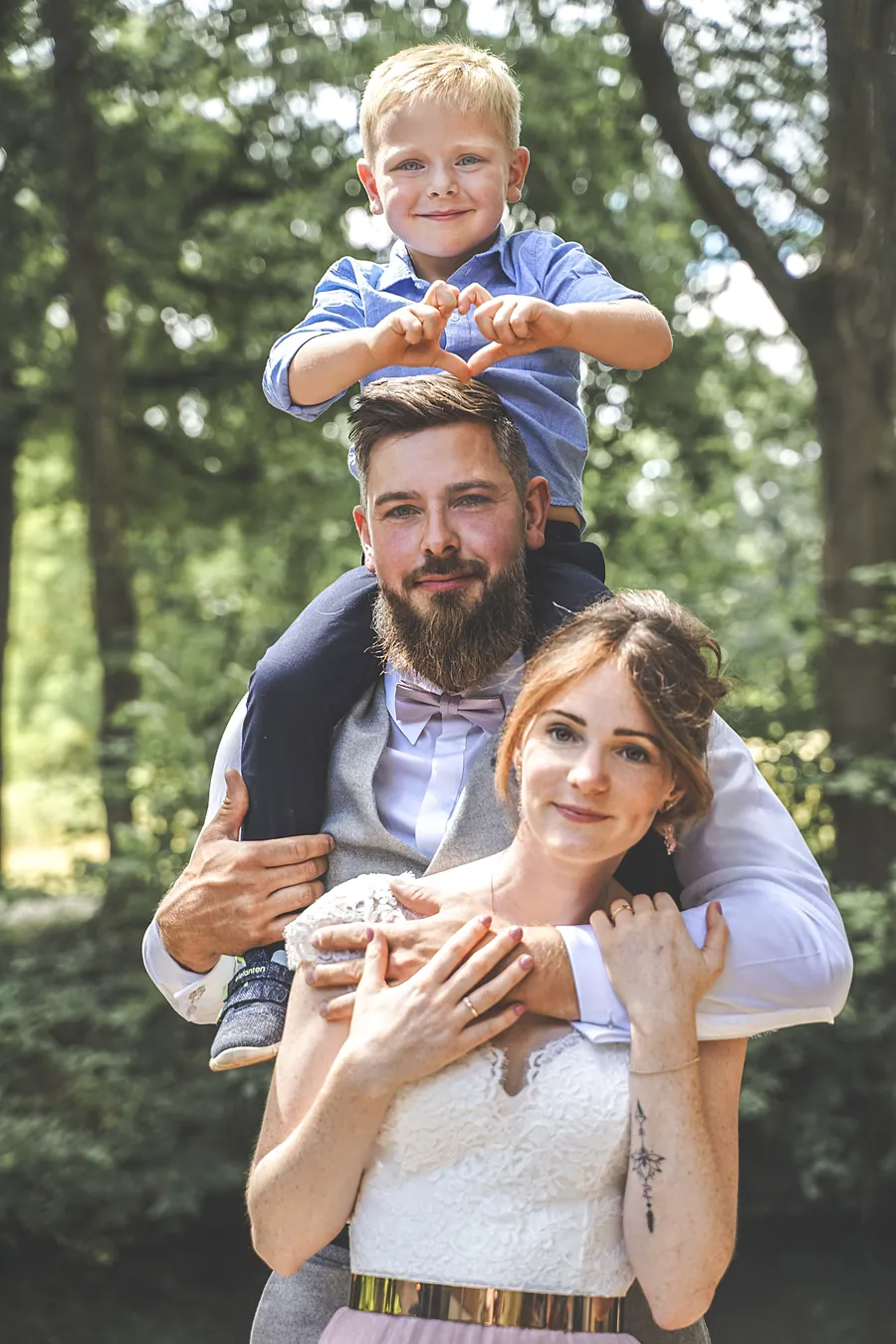 Ein Mann mit einem Jungen auf den Schultern umarmt eine Frau im weißen Kleid im Wald