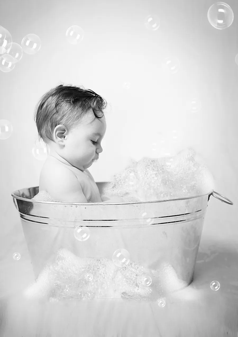 Schwarzweißbild: Ein Baby sitzt in einer kleinen Zinkwanne voller Schaum und schaut Seifenblasen nach