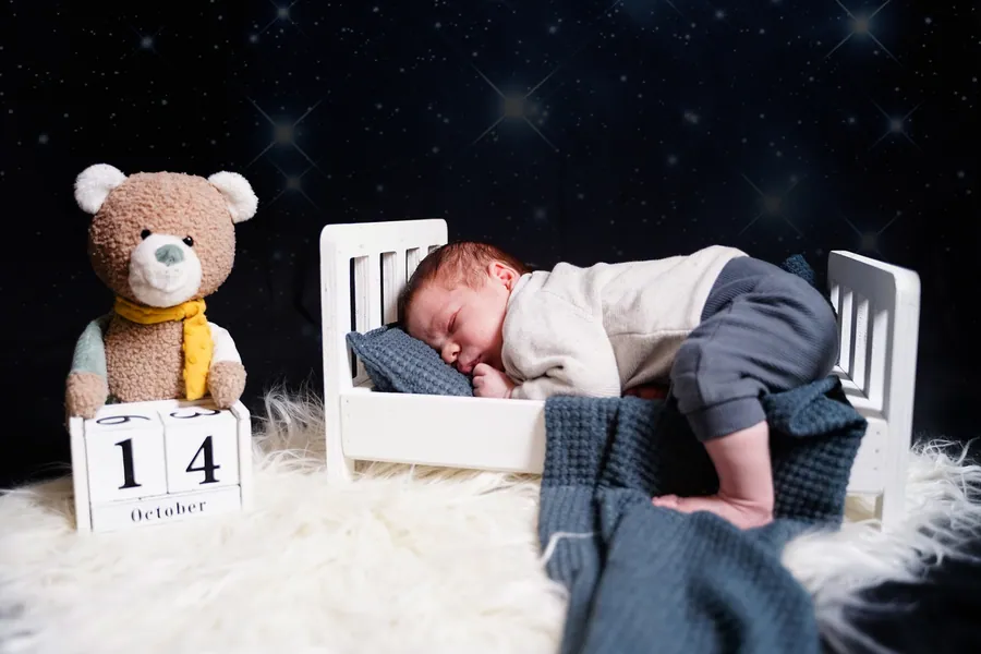 Ein Neugeborenes schläft in einem kleinen weißen Bettchen neben einem Teddy vor sternenklarem Hintergrund