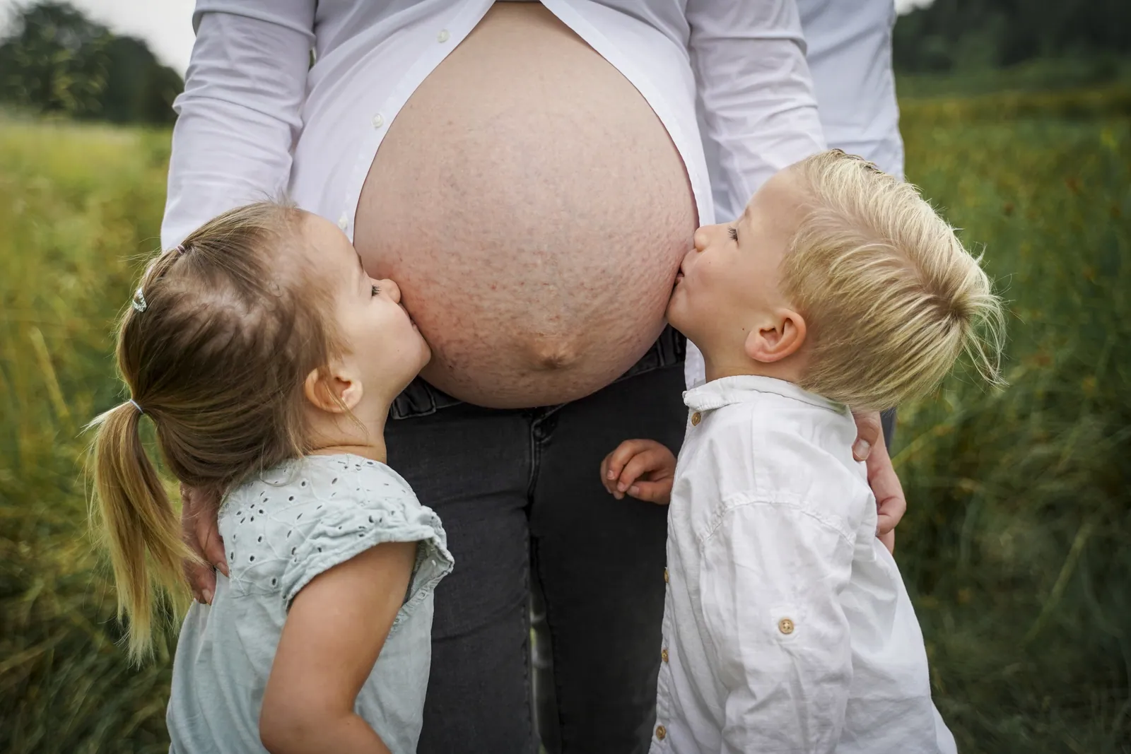 Zwei Kinder küssen den Babybauch ihrer Mutter auf einer Sommerwiese