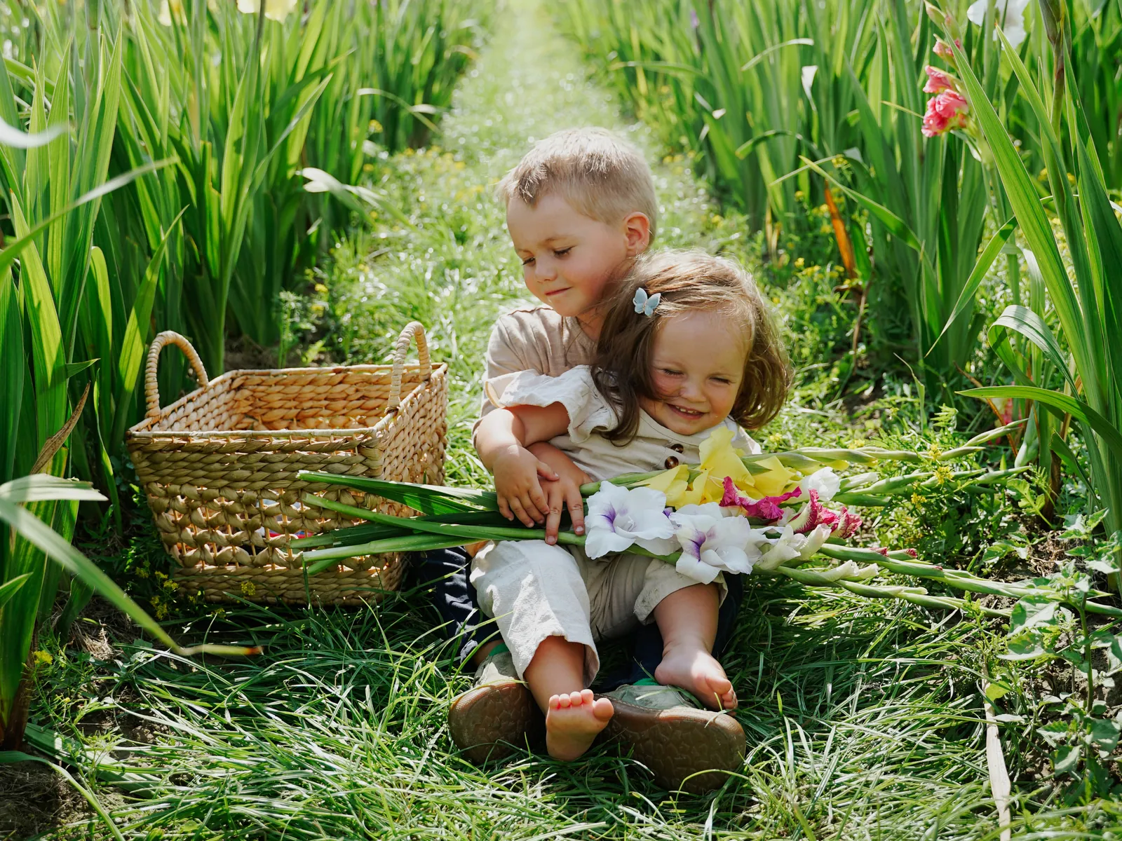 Zwei kleine Kinder sitzen eng beieinander mit einem Picknickkorb und Blumen im hohen Gras