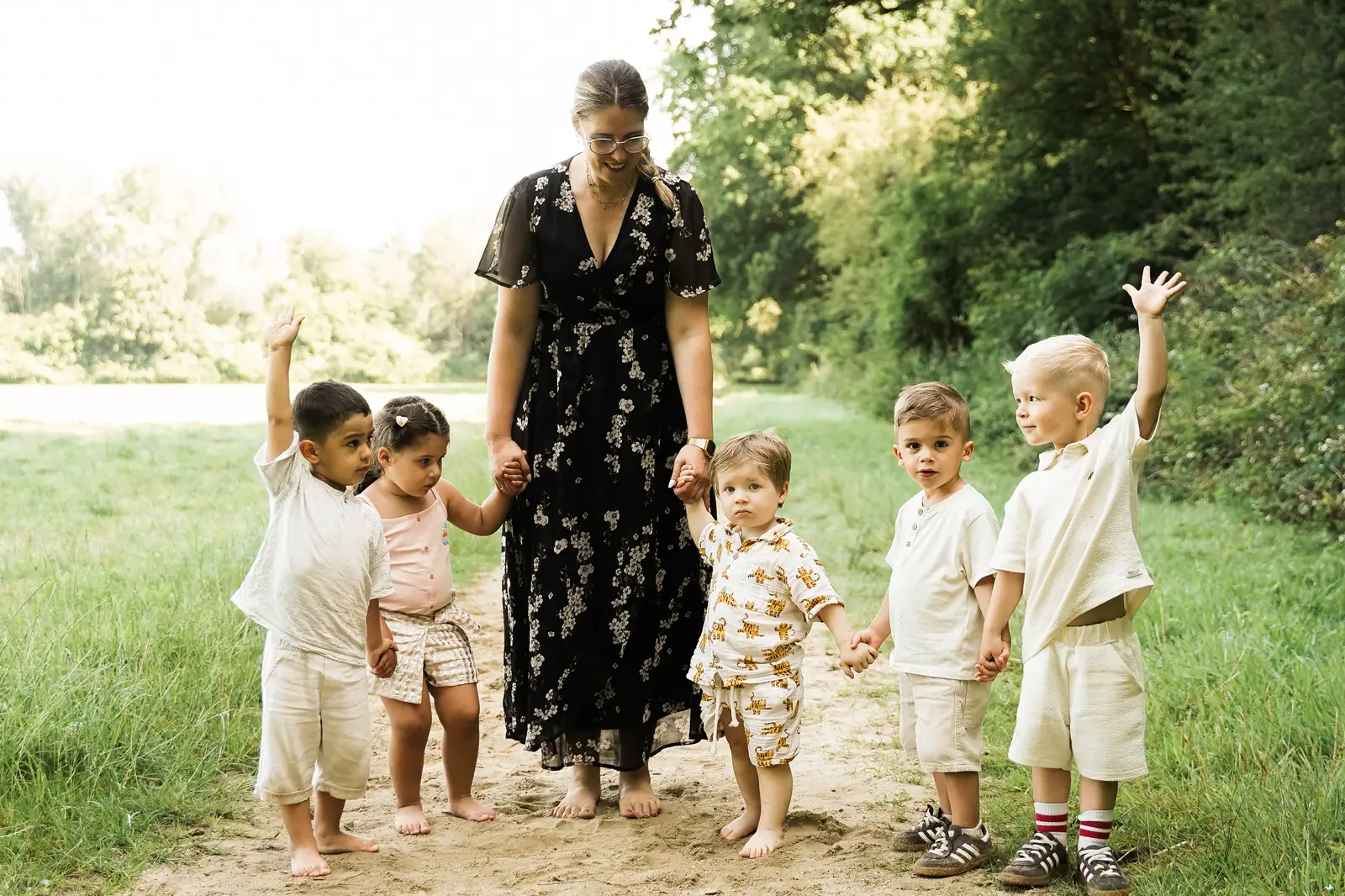 Fünf kleine Kinder laufen Hand in Hand mit einer Erwachsenen über einen sandigen Weg am Waldrand