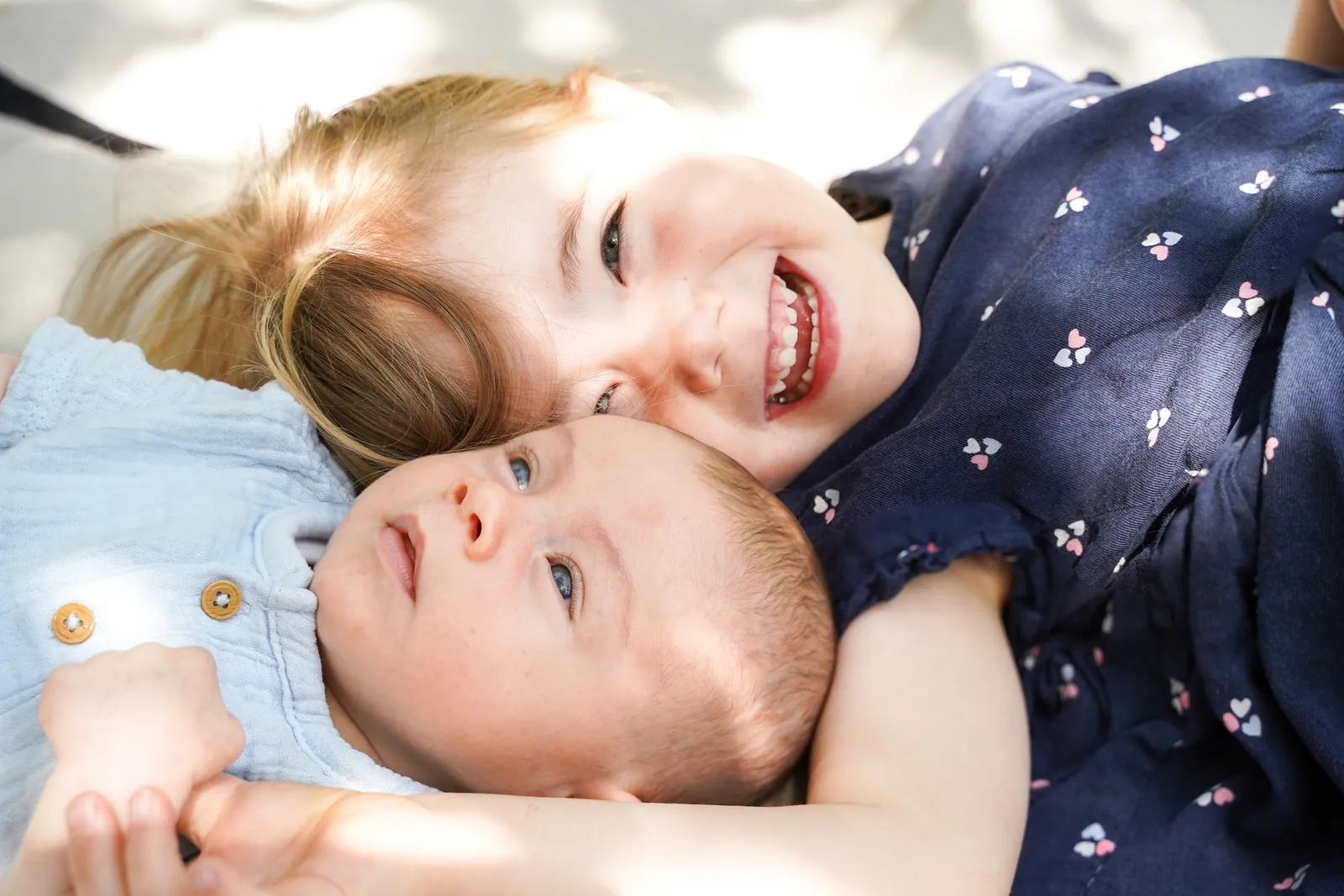 Ein Geschwisterkind kuschelt lachend mit dem Baby im weichen Sonnenlicht zuhause