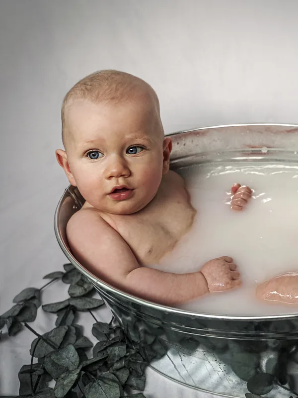 Ein Baby sitzt in einer Zinkwanne mit milchigem Wasser, daneben liegen Eukalyptuszweige